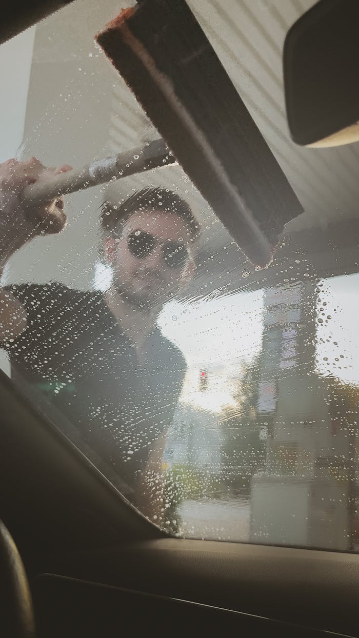A man in sunglasses washes a car window at a gas station using a squeegee.